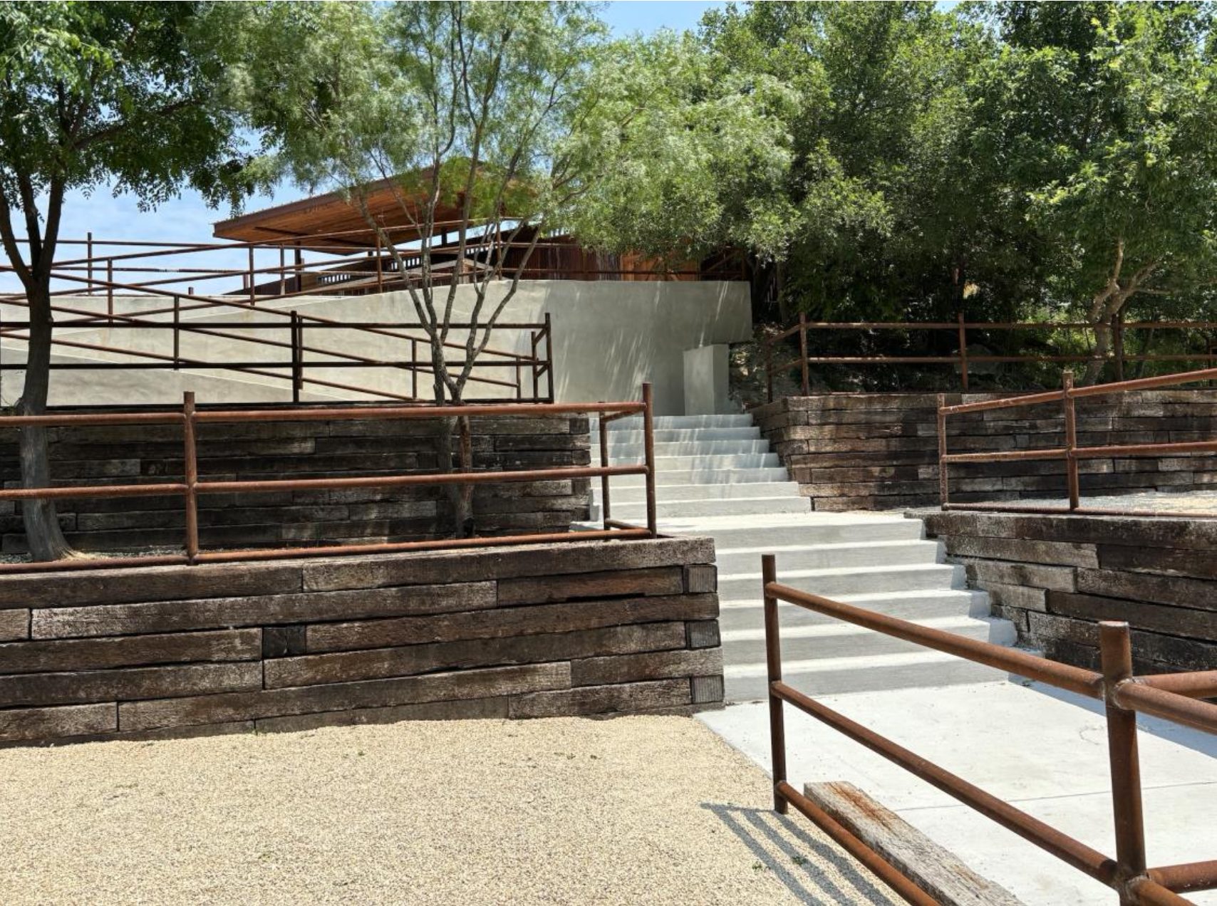 Concrete steps lead up through the tiered amphitheater toward the dancehall, framed by rust-finished railings and cypress trees.